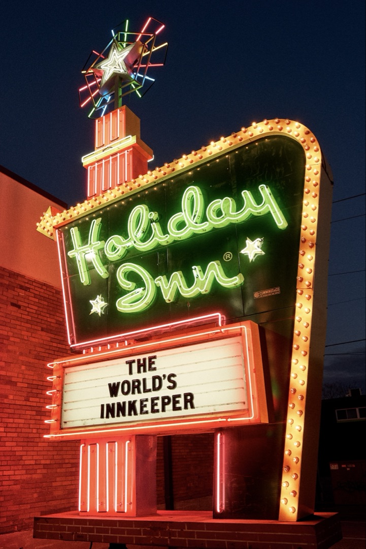The fully restored Holiday Inn Great Sign illuminated at dusk at the American Sign Museum, Cincinnati — its emerald green neon, yellow starburst, and chasing bulbs blazing against the twilight sky
